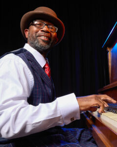 Photo of William Knowles sitting at a beautiful upright piano, dressed as Fats Waller.