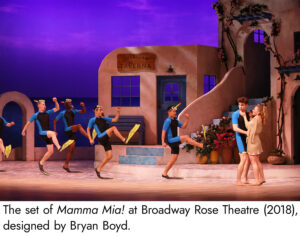 Photo of the set of Mamma Mia, the floor painted to be a sandy beach complete with ripples in the sand to show where the ocean's waves ebb and flow. The buildings up stage are distinctly mediterranean, with a stucco clay look and covered with climbing ivy.