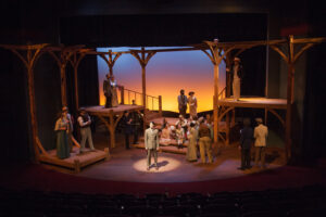 Photo of the set of Parade. Tall wooden beams support wooden platforms of various levels around the stage with a small wooden staircase at center stage. The screen behind the set is lit with vibrant amber at the bottom and grading to a deep blue on top, emulating a sunrise or sunset.