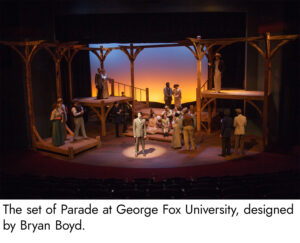 Photo of the set of Parade. Tall wooden beams support wooden platforms of various levels around the stage with a small wooden staircase at center stage. The screen behind the set is lit with vibrant amber at the bottom and grading to a deep blue on top, emulating a sunrise or sunset.