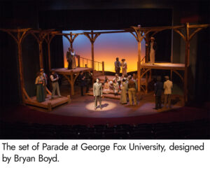 Photo of the set of Parade. Tall wooden beams support wooden platforms of various levels around the stage with a small wooden staircase at center stage. The screen behind the set is lit with vibrant amber at the bottom and grading to a deep blue on top, emulating a sunrise or sunset.