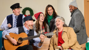 A photo of the cast of Home for the Holidays, sitting together on one of the bus station's benches, joyfully singing a carol.