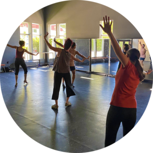 Photo from a 2023 adult dance class showing participants performing a move with their arms outstretched, with the light from the studio windows beaming in from the corner.