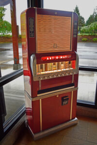 Photo of the Art-o-mat machine in the lobby. It stands about 5 and a half feet tall and is in a midcentury, 1950s industrial chic design. It is a bold cherry red with chrome accents and knobs. A small window in the front that used to show the cigarette brands inside now displays the artists' names and samples of their work.
