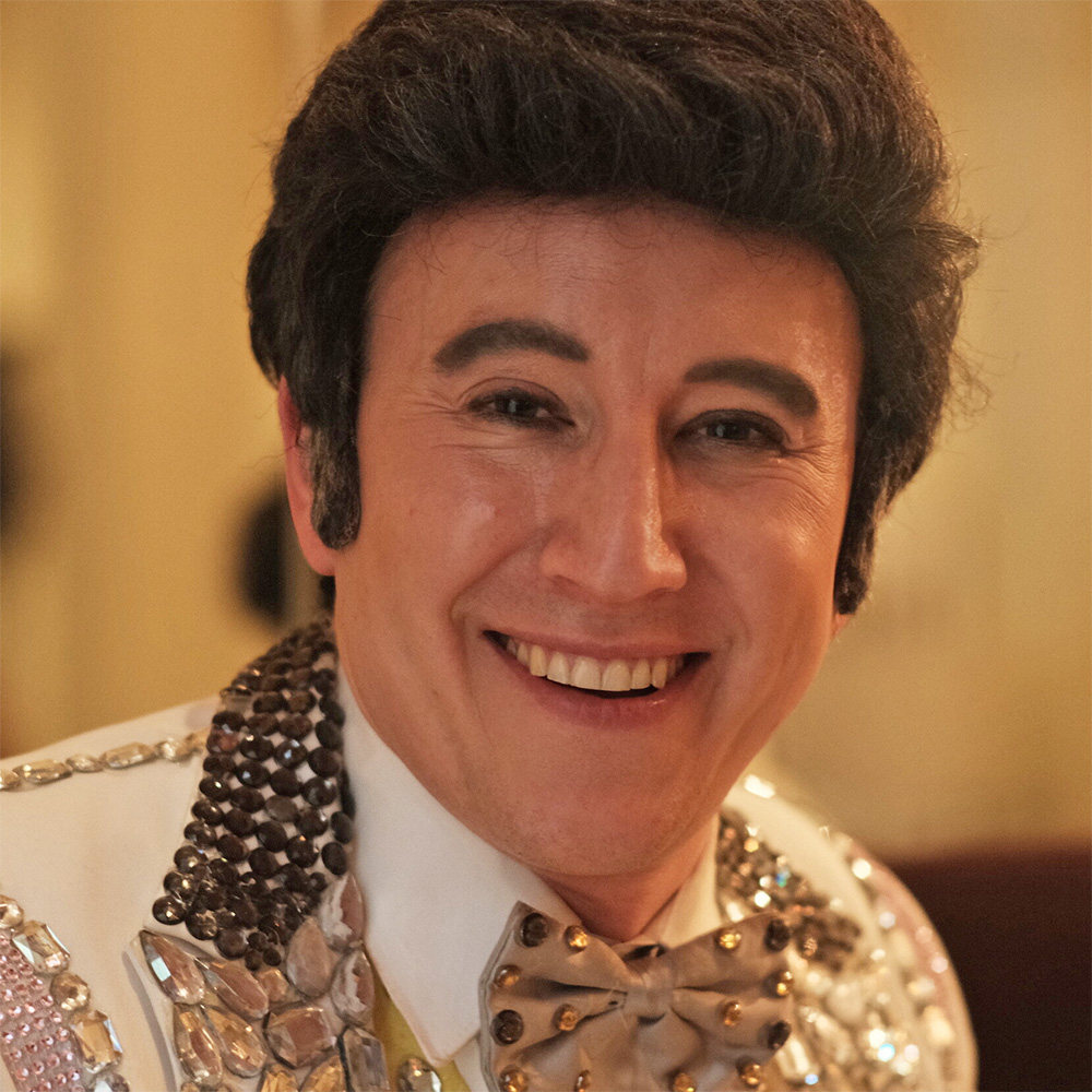 Photo of David Saffert as Liberace sitting in a dimly lit restaurant.