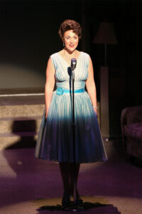 Photo from our 2018 production of Always Patsy Cline with Sara Catherine as Patsy Cline standing in front of an old chrome microphone in a pale blue dress.