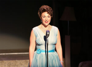 Photo from our 2018 production of Always Patsy Cline with Sara Catherine as Patsy Cline standing in front of an old chrome microphone in a pale blue dress.