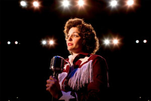 Photo of Sara Catherine Holder as Patsy Cline. She is lit with a soft amber light and standing alone on a stage, holding an old, chrome microphone.
