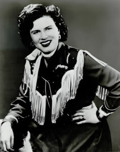 Promotional photo of Patsy Cline, posing in her iconic cowgirl outfit with white fringe.