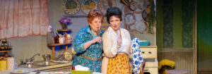 Photo of Sharon Maroney and Sara Catherine Holder in Always Patsy Cline 2024 production. They are standing in Louise's (Sharon's) kitchen, singing with wooden spoons for microphones.