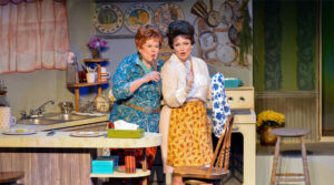 Photo of Sharon Maroney and Sara Catherine Holder in Always Patsy Cline 2024 production. They are standing in Louise's (Sharon's) kitchen, singing with wooden spoons for microphones.