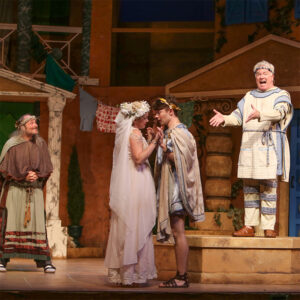 Photo from our production of A Funny Thing Happened on the Way to the Forum. Dan Murphy is standing on a box mid stage narrating the action around him as two lovers kiss and an disheveled man looks on.