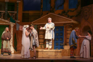 Photo from our production of A Funny Thing Happened on the Way to the Forum. Dan Murphy is standing on a box mid stage narrating the action around him as two lovers kiss and an disheveled man looks on.