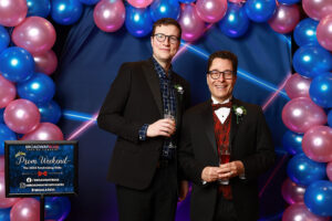 Photo of guests at the prom photo booth at the event. They are in front of a blue and pink balloon arch and blue and pink 1990s themed backdrop.