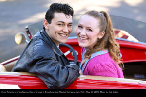 Photo of Victor Polanco and Sophie MacKay in a beautiful old, red sports car. They are turned around, looking backward toward the camera.