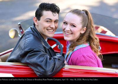 Photo of Victor Polanco and Sophie MacKay in a beautiful old, red sports car. They are turned around, looking backward toward the camera.