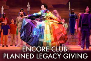 Photo from our 2023 production of Joseph and the Amazing Technicolor Dreamcoat. Shows Joseph holding out his coat of many colors proudly. Text overlaid reads Encore Club, Planned Legacy Giving.