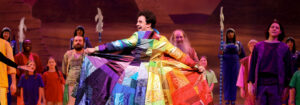 Photo from our 2023 production of Joseph and the Amazing Technicolor Dreamcoat. Shows Joseph holding out his coat of many colors proudly.