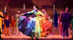 Photo from our 2023 production of Joseph and the Amazing Technicolor Dreamcoat. Shows Joseph holding out his coat of many colors proudly.