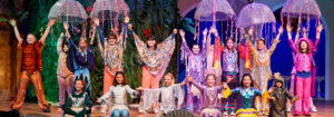 Photo from the 2025 kids production of Little Mermaid, showing the children's ensemble spread across the stage with their arms open wide at the end of a song. They are dressed as fish, jellyfish, and other cute sea animals.