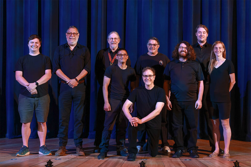 Photo of the Anything Goes band. They are all on the front of the stage with the blue curtain behind them. They are dressed in black, about to play for their first live audience.