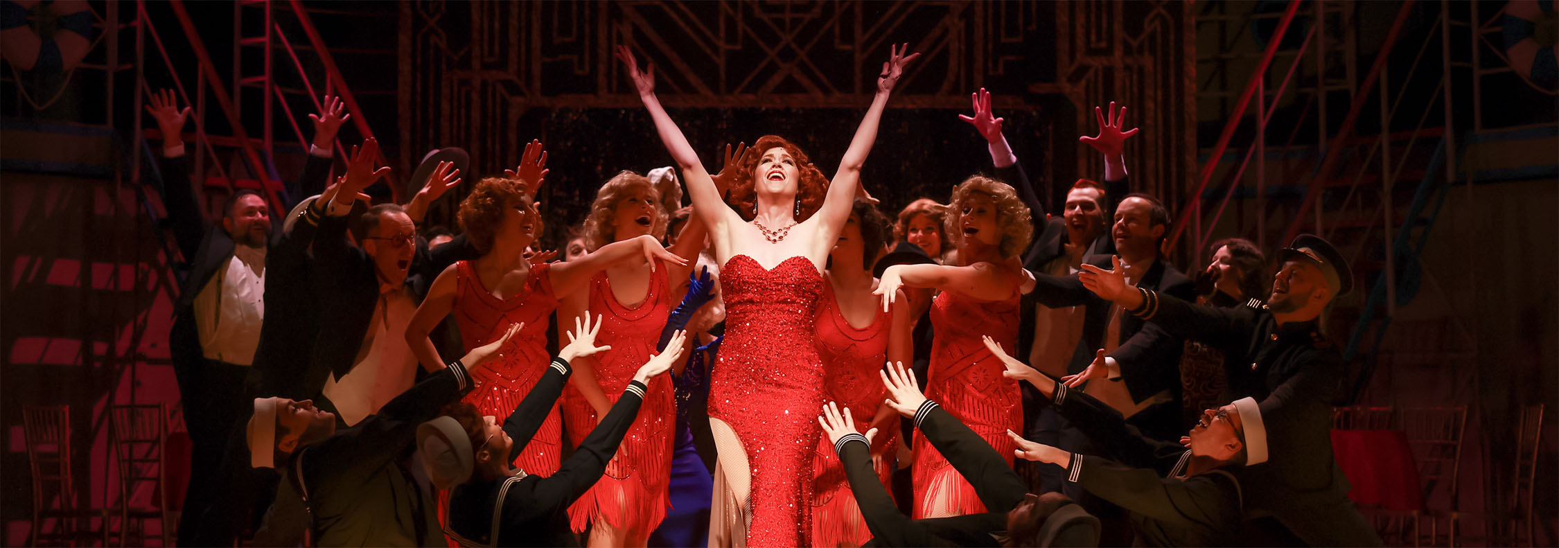 Photo from Anything Goes of the end of the song Blow Gabriel Blow. The entire cast is crouch and kneeling around Reno Sweeney, the nightclub singer, as she is posing with both of her hands in the air. She is wearing a sparkling, strapless red gown and the stage is bathed in red light, while Reno in the middle has a bright spotlight.