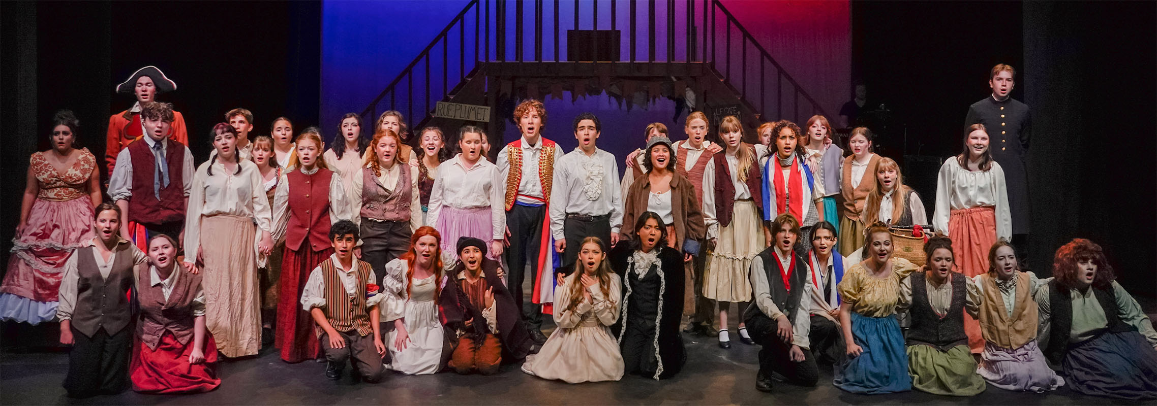 Photo of the cast of Les Misérables School Edition singing out to the audience. The whole cast is spread across the stage, singing together at the end of the show.