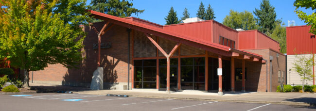 Photo of the entrance of the Broadway Rose New Stage. It is late summer in this picture, with a bright blue sky and the tops of the nearby trees beginning to turn red and orange. The building has an angled roof awning over the main entrance. The building is red brick on the lower half and red-painted corrugated metal on the upper half and on the roof.