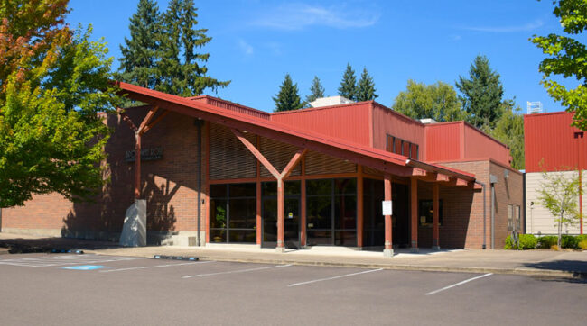 Photo of the entrance of the Broadway Rose New Stage. It is late summer in this picture, with a bright blue sky and the tops of the nearby trees beginning to turn red and orange. The building has an angled roof awning over the main entrance. The building is red brick on the lower half and red-painted corrugated metal on the upper half and on the roof.