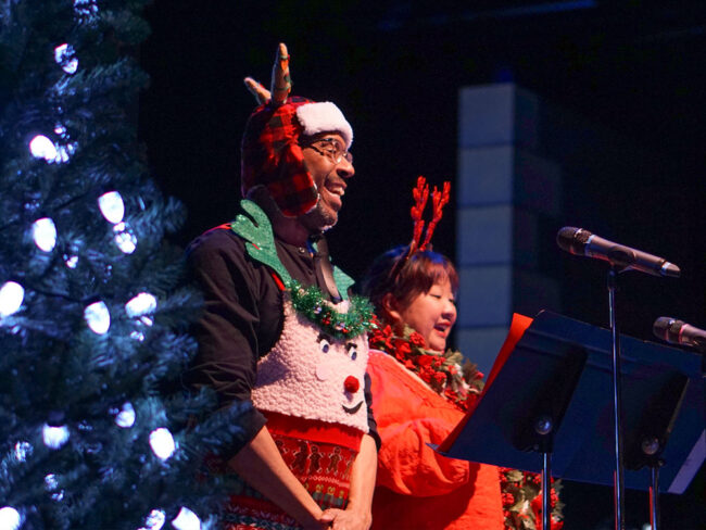 Photo from one of our Santa Sing-Along events for families the first weekend of December. This photo shows two of our guild members up on stage in festive costumes leading a sing-along with an audience full of families.