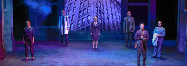 Photo from Triangle of the beginning of the show with the full cast spread out across a blue-tinted, dimly lit stage. They are all staring out into the audience, looking slightly up. Behind them is a very large image of the building where the real life Triangle Shirtwaist Factory Fire took place. A haze is filling the stage.