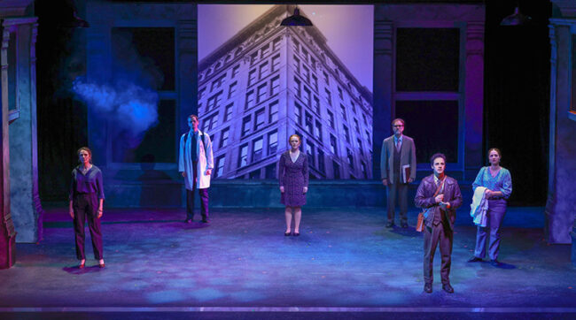 Photo from Triangle of the beginning of the show with the full cast spread out across a blue-tinted, dimly lit stage. They are all staring out into the audience, looking slightly up. Behind them is a very large image of the building where the real life Triangle Shirtwaist Factory Fire took place. A haze is filling the stage.