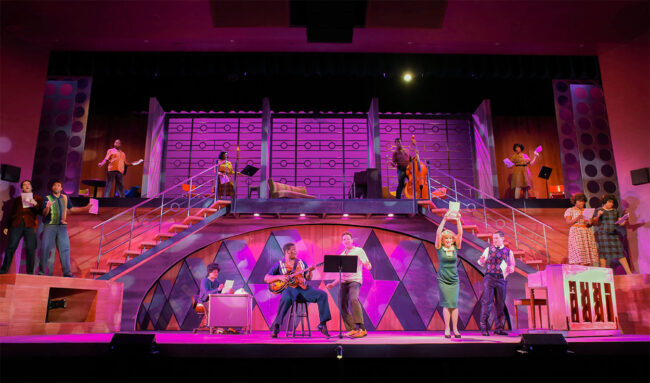 Photo from Beautiful, the Carole King Musical. Shows a wide shot of the whole stage as the song 1650 Broadway Medley is performed by the majority of the cast spread across the stage. The set is in multiple levels with large staircases on either end of the stage leading up to a high catwalk platform making a second floor for the set. The cast is bathed in pink light as the dance and sing a medley of 1950s pop and soul hits.