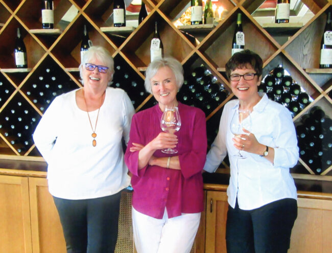 A photo with Heidi Heydlauff in the middle, director Lyn Cramer on the right, and Heidi's good friend Carrie Reynolds on the left. They are standing and smiling for the camera at the tasting room of a winery, a wall of wine bottles is behind them.