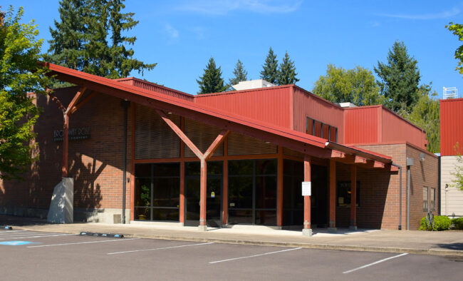Photo of the entrance of the Broadway Rose New Stage. It is late summer in this picture, with a bright blue sky and the tops of the nearby trees beginning to turn red and orange. The building has an angled roof awning over the main entrance. The building is red brick on the lower half and red-painted corrugated metal on the upper half and on the roof.