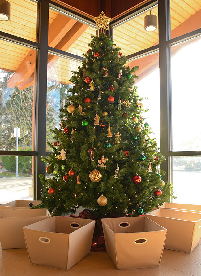 A photo of the Christmas tree in our lobby with fabric bins beneath it, ready to be filled by our patrons generous donations to the poor and houseless.