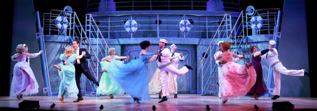 A photo from our recent production of Anything Goes during the starlight dancing scene with couples dressed in pastel colored ball gowns and sailor uniforms. All the dancers are mid-leap during the photo with the women's dresses elegantly wafting around from the dramatic move.