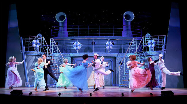 A photo from our recent production of Anything Goes during the starlight dancing scene with couples dressed in pastel colored ball gowns and sailor uniforms. All the dancers are mid-leap during the photo with the women's dresses elegantly wafting around from the dramatic move.