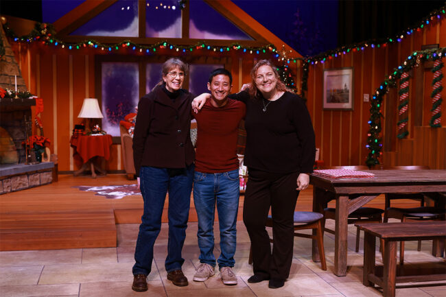A photo of our band members Katie Nordone on bass (left), Yuya Matsuda on drums (center), and music director Wendy Vece on keys (right). They are standing on the show's set with all the colorful lights on. They have thier arms over each other's shoulders.