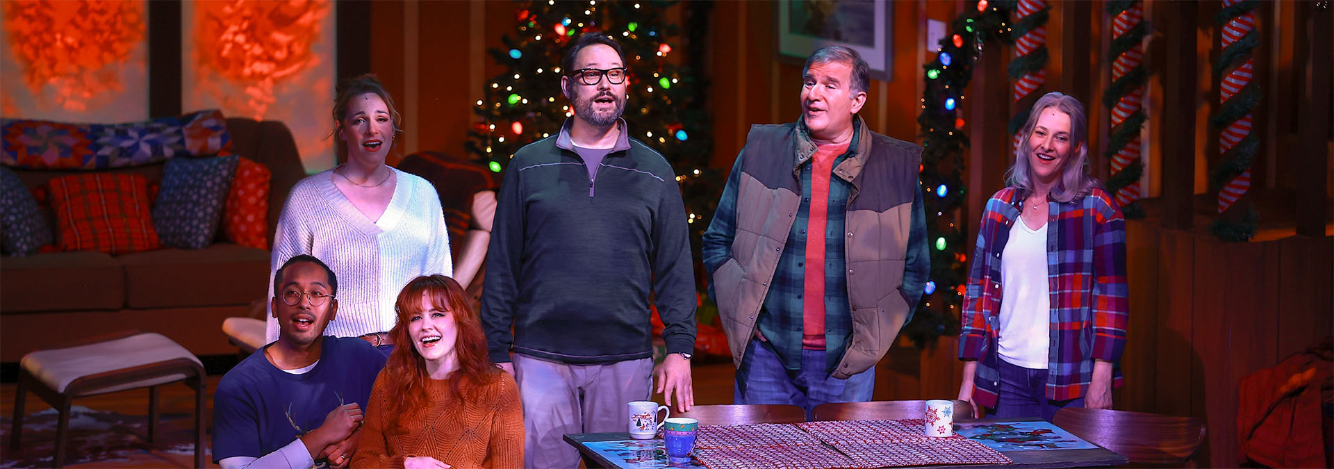 A photo from Christmas in Door County showing all 6 cast members standing and sitting around the dining table. They are all singing out to the audience, the lights are low, but the Christmas tree lights a twinkling colorfully behind them.
