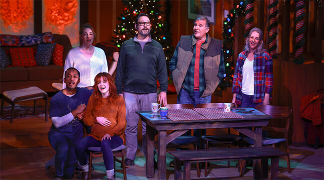 A photo from Christmas in Door County showing all 6 cast members standing and sitting around the dining table. They are all singing out to the audience, the lights are low, but the Christmas tree lights a twinkling colorfully behind them.
