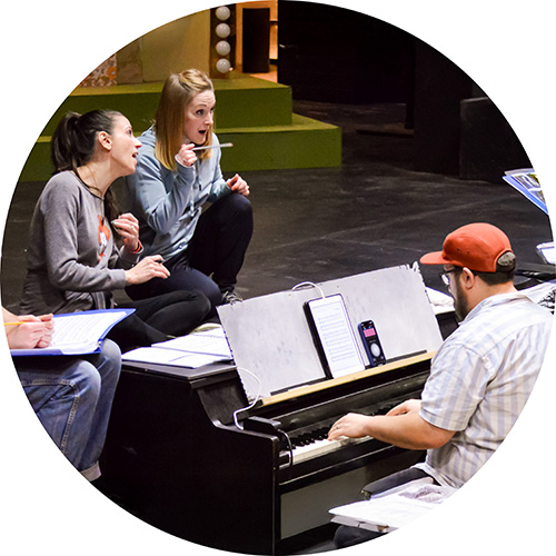 Photo of 3 performers on a stage sitting and crouched around a piano sitting on the floor in front of the stage. They are all singing together, concentrating.