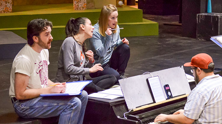 Musical Theatre Scene Study Photo of 3 performers on a stage sitting and crouched around a piano sitting on the floor in front of the stage. They are all singing together, concentrating.
