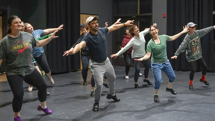 Tap - Advanced A photo from one of our advanced adult tap classes showing the students doing a big broad move with their arms outstretched and leaping onto one foot.
