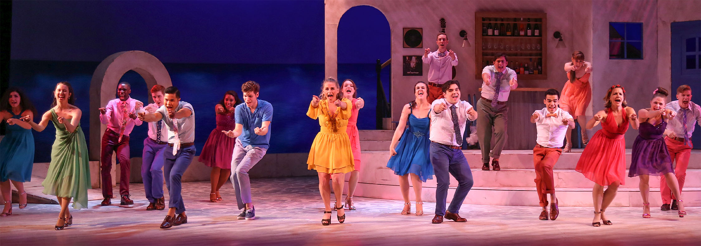 A photo of the cast of Mamma Mia in a line across the stage, all dancing and singing out to the audience with smiles and brightly colored, splashy costumes.