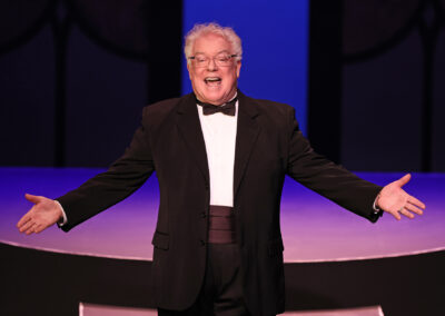 A photo of Dan Murphy in a tuxedo hosting a recent Broadway Rose fundraiser gala. He is smiling big while he speaks and holding his arms out wide in a welcoming gesture.