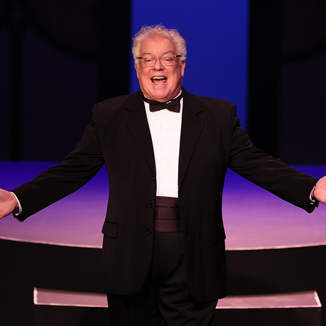 A photo of Dan Murphy on stage hosting a gala in a tuxedo, holding hims arms out in a grand gensture.