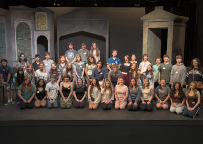 Photo of the entire Teen Summer Production cast. Roughly 40 teens are spread across the stage, all smiling for the camera. They are on the beginnings of the Hadestown set, with old stone structures like cemetery buildings and mausoleums behind them.