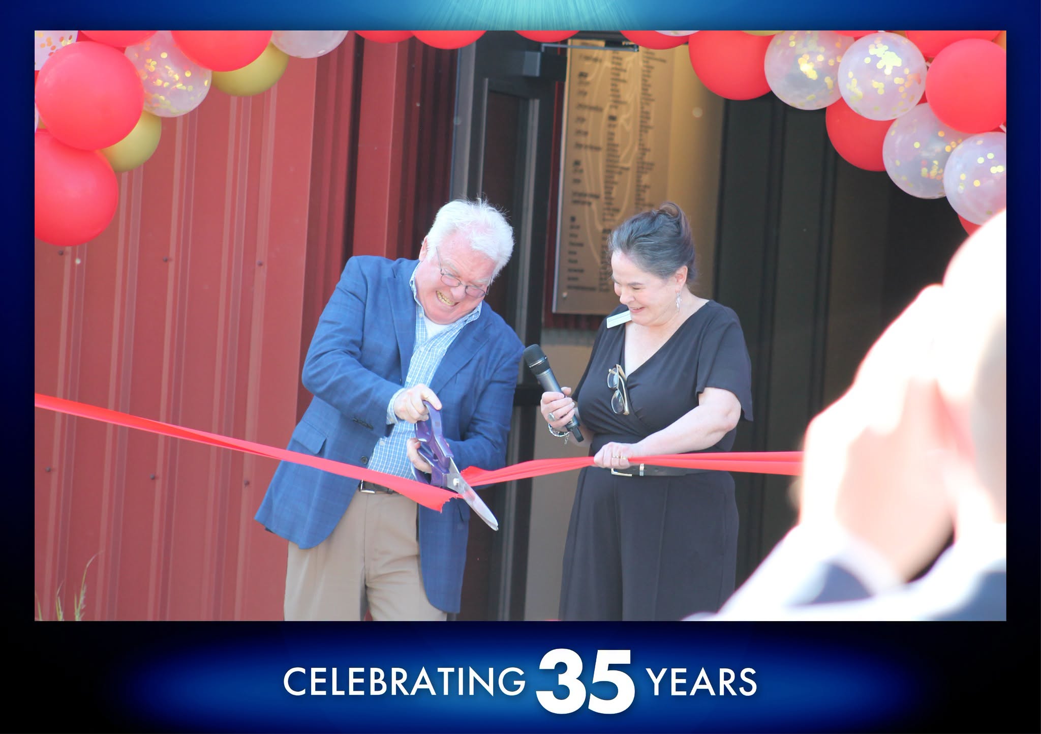 Dan and Sharon cut the ribbon A photo of Dan Murphy trying to cut a red ribbon with a large pair of scissors in front of the Broadway Rose Theatre's new addition in 2022. Sharon Maroney, his wife and Broadway Rose Artistic Director, looks on amused at his struggle.