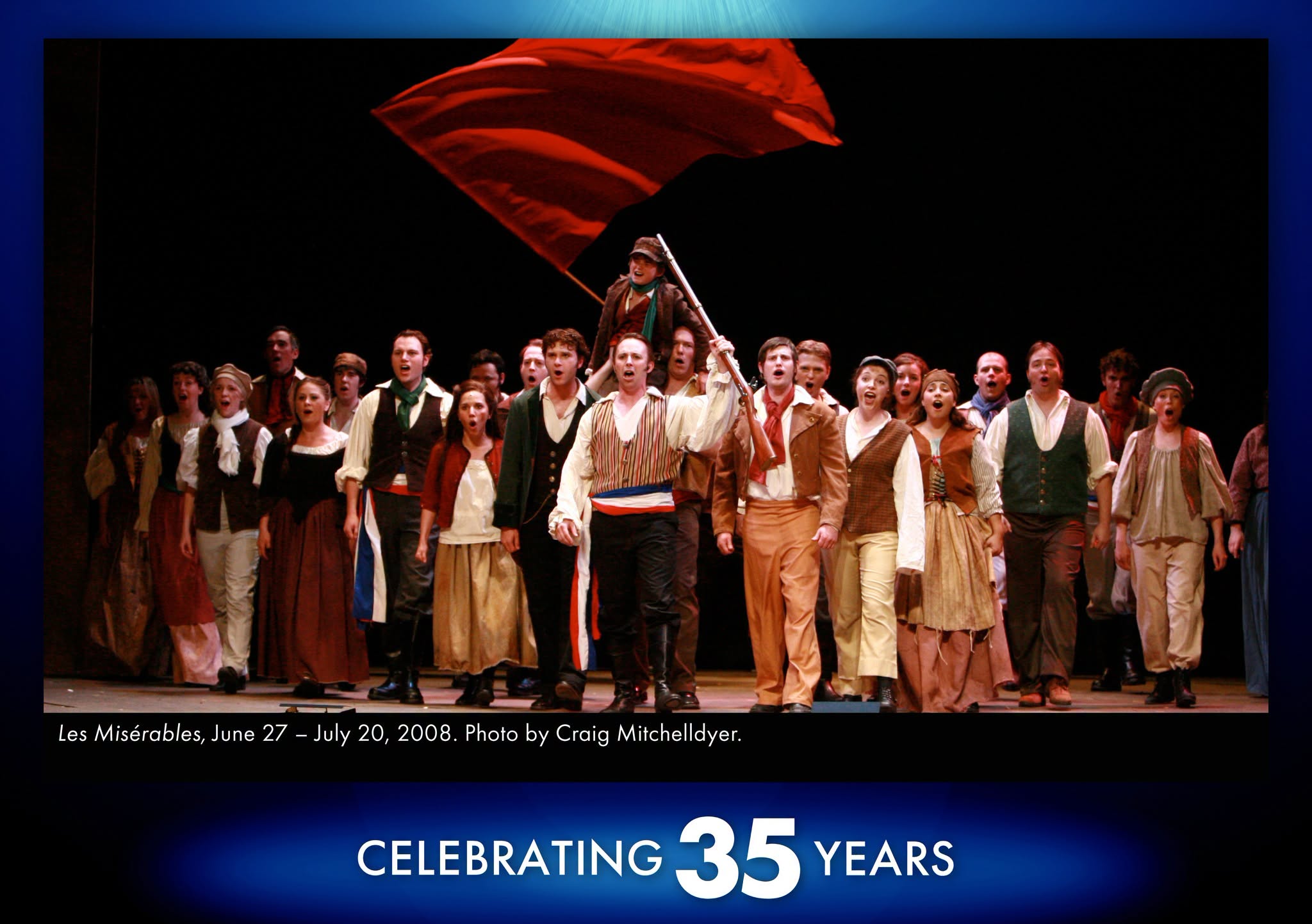 Les Miserables A photo from our production of Les Miserables that shows the cast in a large group singing together passionately with a large red flag waving behind them as the characters prepare for a revolution.